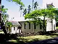 Photograph of the church established by Father Damien at the Kalaupapa Leprosy Settlement, behind a low stone wall and surrounded by palm and other trees.
