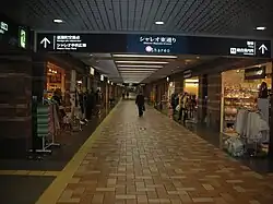Long corridor with an illuminated electric sign at the overhead entrance. Open stores line either side of the corridor.