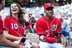 Miguel Batista (right) and Katherine Connors both wearing red Washington Nationals jerseys, holding microphones, and laughing during an interview at Nationals Park