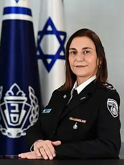 Katy Perry, a uniformed Israeli police officer, sits at a table with her hands folded, posing in front of an Israeli flag and the flag of the Israel Prison Service.