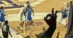 Basketball players watch a ball rebound while fans hold up an "ok" hand sign with three fingers pointed upwards in the foreground.