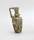 Jug that is pale green glass with bluish-grey tinge; mould-blown, trail-decorated and tooled. Decoration consists of a row of six figures with prominent headdresses or haloes, one on each facet of the body. The figures are represented in frontal view, standing with their arms by their sides, each holding an object. They are dressed in knee-length garments, their legs somewhat apart and their feet shown from the side. Some of the figures may be musicians – two hold what appear to be flutes – but the remaining objects are not clear enough to be identifiable
