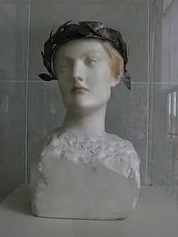 A marble bust of a woman with a calm, distant expression wearing a dark laurel wreath, the shoulders roughly carved compared to the smooth face