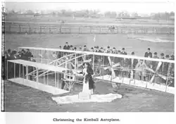 A biplane with multiple propellers sitting on the ground. Around it are approximately 40 men. A woman in fancy clothes is standing in front of the plane.