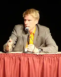 Christof Koch wearing a yellow t-shirt under a yellow shirt, with a dark blazer, holding a laptop computer in his left hand, grinning at camera