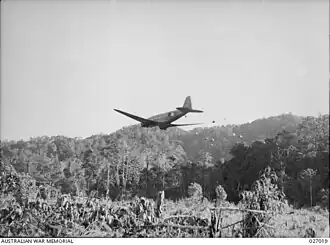 A transport plane flying at low level away from the camera, dropping supplies over a clearing in the jungle