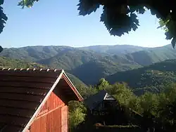 Red house in the left foreground. The rest of the picture depicts lush green mountainous hills and valleys of Serbia.
