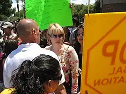 A woman in her thirties with fairly short blond hair, wearing sunglasses and a beige and pink top, is surrounded by a crowd.