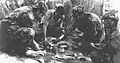 Portuguese troops eating rations on the cloth of a tent laid out on the ground.