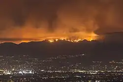 Fire and smoke glow orange at the top of dark hills, with a field of city lights far below between the photographer and the hills