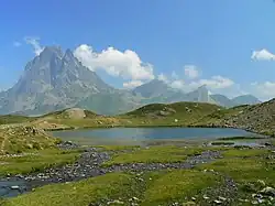 Colour photograph showing several streams flowing into a lake surrounded by meadows and scree. In the background, a rocky peak dominates the landscape.