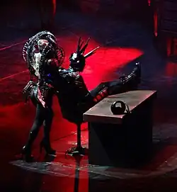 A woman, sporting a ram-horned headpiece, stands behind a man sitting in a chair.