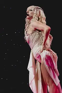 Lady Gaga singing while wearing a platinum blonde wig, an ivory outfit, and a headset microphone