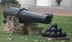 A long 19th-century cannon with a stack of cannonballs in front of it, sitting on a lush green lawn.
