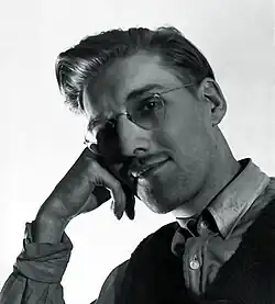Black-and-white photograph of a man in glasses resting his head on his hand and smiling at the camera