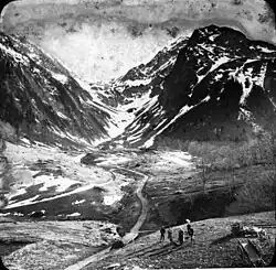 Black and white photo, a group of five people stand near a path that enventually winds between two snow covered peaks.