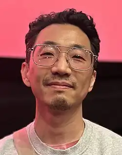 A man wearing glasses and looking at the camera with a slight smile. The background is split pink and black.