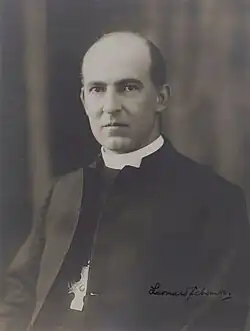 Frowning, balding man in forties seated in clerical dress and cross necklace