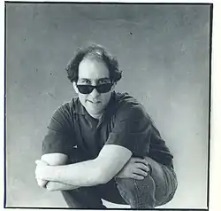 Man crouching in a studio wearing dark sunglasses, a short-sleeve polo shirt, and jeans, with thinning hair on top and fuller, wavy hair at the sides
