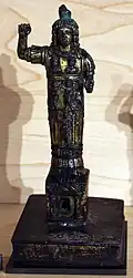 Photograph of an ancient bronze statue of a god with a raised right hand, ornate robe, and intricate details on a pedestal, set against a neutral background.