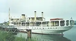 A white ferry with several large windows around the outside sits in water next to a jetty.