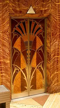 Ornate elevator doors with Art Deco design of stylized papyrus plants in gold and brown against marbled stone wall