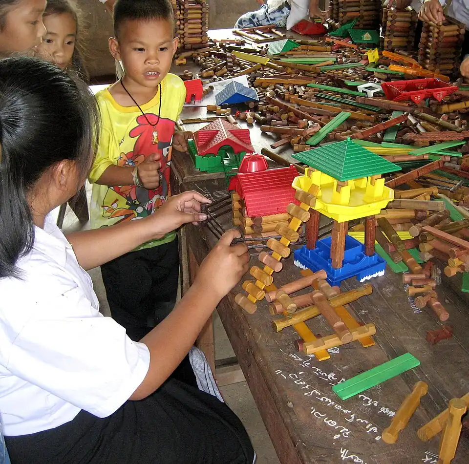 A photo of wooden table strewn with Lincoln logs. In the very foreground, there is chalk writing on the table, in Lao script. Further back there are models of a stilt house and single-storey houses (the latter enclosed by fences).