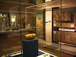 View of the Cyrus Cylinder in its display cabinet, situated behind glass on a display stand. Other ancient Persian artefacts can be seen lining the room in the background.