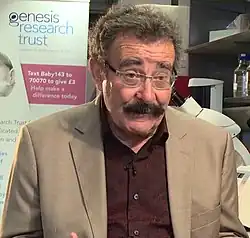Robert Winston, wearing a burgundy-coloured, patterned shirt, under a tan jacket, looking just down and left of camera, appearing to speak to someone off-camera. In the background there are various paraphernalia suggesting he may be in a science laboratory.