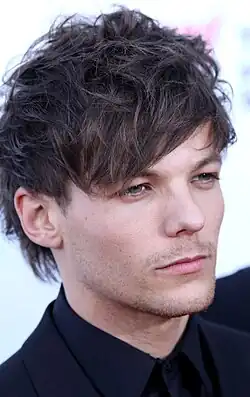 A headshot of a man in his early 20s. A black suit, shirt, and tie are visible. He has blue eyes and messy brown hair.