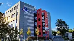 A modern multi-story building with a gray panel façade and a vertical red section featuring large circular cutouts, surrounded by trees and a pedestrian sign, with a blue sky in the background.