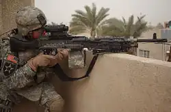 Soldier in combat gear holding an M240B machine gun fitted with an M145 machine gun optic, positioned over a balcony.
