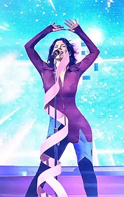 A woman perfoming onstage, wearing a purple jumpsuit with two blue stars over the knees, showcasing a starry background.