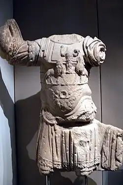 Photograph of an ancient stone torso sculpture of a headless figure in detailed armor. The piece displays intricate carvings and conveys a sense of historical grandeur.