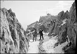 Two men stand between two rock faces with a stone foundation behind them in the distance. They are facing the direction of the camera looking right. The man on the right is pointing upwards off camera.