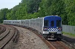An eight-car passenger train, silver on the sides and blue on the front, on a curved stretch of track with a third rail