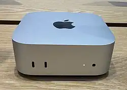 A Mac mini computer (shaped like a small aluminum box) is pictured on top of a wooden table.