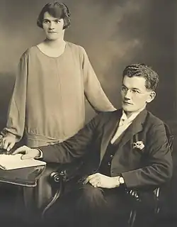 "Greyscale photograph showing a woman in a dress and with a fashionable hairstyle; she is standing to the right of and slightly behind a bespectacled man wearing a suit and breasted jackey seated on a chair with his right hand touching a white book on a table; each person is looking a little to their right