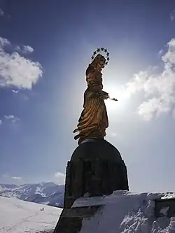 The golden statue of Our Lady, Protector of Europe created by Casagrande, standing on a stone pedestal with the sun behind her head illuminating her twelve-star halo, with snow-covered mountains in the distance