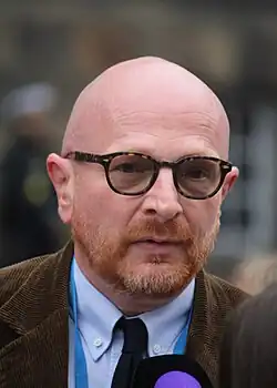 close-up of Mads Brügger wearing a light blue shirt, brown suit jacket, and dark tie, looking at camera