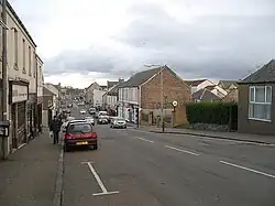 A view down Kelty's Main Street, lined by a number of small shops. There are several parked cars, and one man with a carrier bag walking up the pavement.