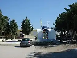 A white building with a blue dome and a flagpole next to trees and a parked car.