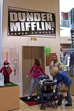 Three adults and a child in a stroller in front of a set of closed elevator doors. Above them is a sign with "Dunder Mifflin Paper Company" on it in white letters on a black background