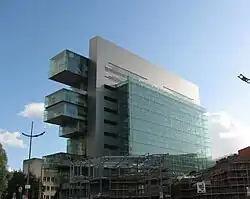 A very modern glass-and-steel large building as viewed from a short distance away against a blue sky. The building is made of a series of connected cuboids, of which only one touches the ground and the others all come off one another.