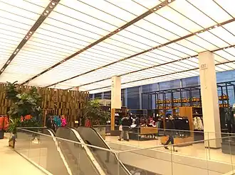 View of the second floor. There are racks of merchandise in the background and an escalator in the foreground. The ceiling is composed of a luminous material, and the lights are hidden behind the ceiling. The glass facade is visible at far right.