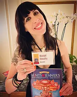 Douaihy posing with ID card and a catalog. She has shoulder-length brown hair and bangs, and wears a low cut sleeveless black top.