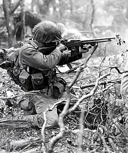 U.S. Marine Corps infantryman kneeling in underbrush and firing a Browning Automatic Rifle while aiming into area out of frame.