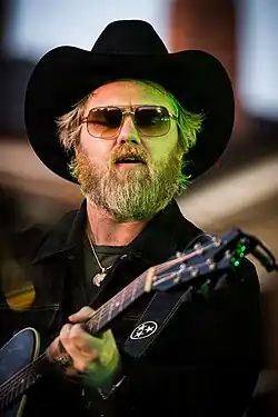 A bearded blond man in a black jacket and black cowboy hat, playing guitar and singing into microphone