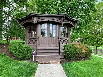 A small, octagonal wooden building with a large porch and a conical roof, nestled among trees.