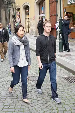 Mark Zuckerberg and his wife Priscilla Chan walking on a cobble stone street in May 2013. They are both wearing jeans. Chan is wearing a scarf and a grey sports jacket. Zuckerberg is wearing a dark grey hoodie. They are both looking at the camera, unsmiling.